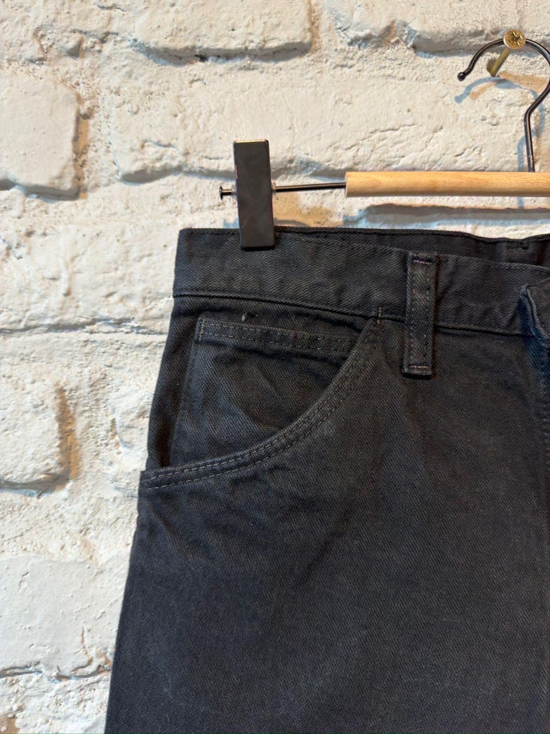 K&ouml;nnte beinhalten: Nahaufnahme einer dunkelgrauen Jeanshose, die an einem Holzb&uuml;gel h&auml;ngt. Die Hose hat ein klassisches F&uuml;nf-Taschen-Design mit einer sichtbaren Vordertasche und G&uuml;rtelschlaufen. Der Stoff scheint ein robustes, strapazierf&auml;higes Material zu sein.