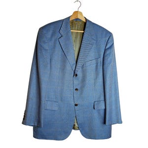 Puede incluir: Una blazer azul claro con un sutil estampado de cuadros, colgada de una percha de madera. La chaqueta tiene un cierre clásico de dos botones, solapas con muescas y dos bolsillos delanteros. El forro interior es de color verde claro.