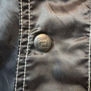 Op de afbeelding: Close-up van een zwarte jas met een knoop met het Fila-logo. De jas heeft witte stiksels langs de naden. De knoop staat in het midden van het frame, met de stof licht gekreukeld.