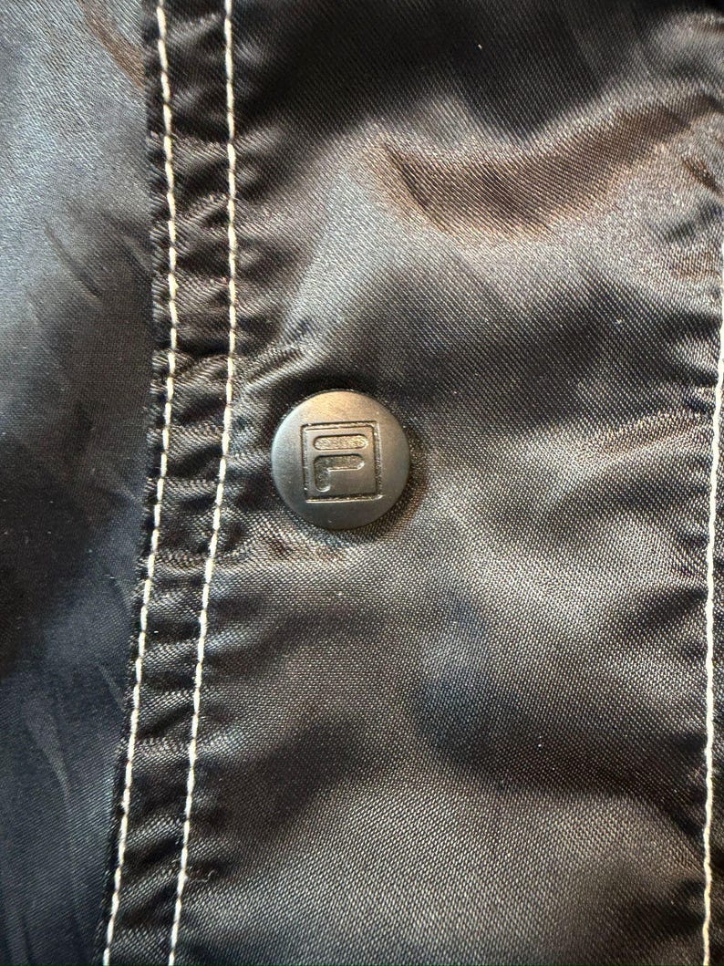 Op de afbeelding: Close-up van een zwarte jas met een knoop met het Fila-logo. De jas heeft witte stiksels langs de naden. De knoop staat in het midden van het frame, met de stof licht gekreukeld.
