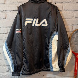 Op de afbeelding: Zwarte Fila-jas met witte en lichtblauwe details. De jas heeft het witte Fila-logo op de rug en een kleine patch op de linkermouw. Voorzien van een hoge kraag en geribde manchetten.