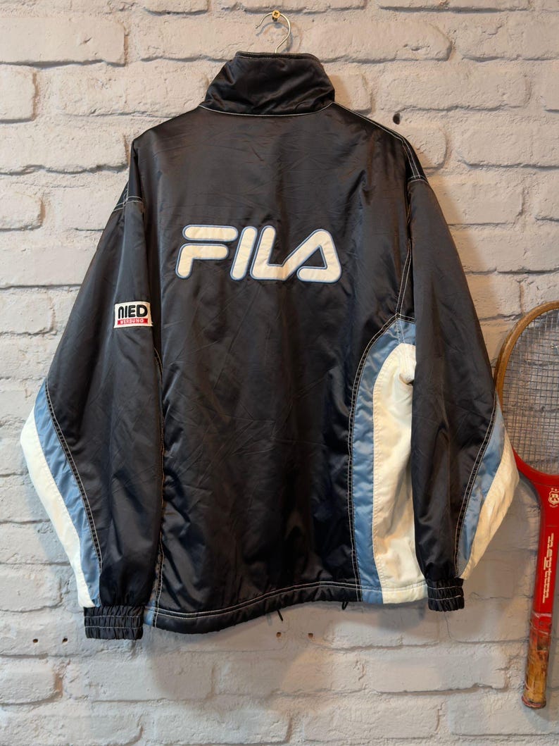 Op de afbeelding: Zwarte Fila-jas met witte en lichtblauwe details. De jas heeft het witte Fila-logo op de rug en een kleine patch op de linkermouw. Voorzien van een hoge kraag en geribde manchetten.