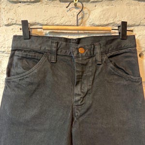 K&ouml;nnte beinhalten: Nahaufnahme einer anthrazitfarbenen Jeanshose, die an einem Holzb&uuml;gel h&auml;ngt. Die Hose hat einen Knopfverschluss und Vordertaschen. Der Hintergrund ist eine wei&szlig;e Backsteinmauer.