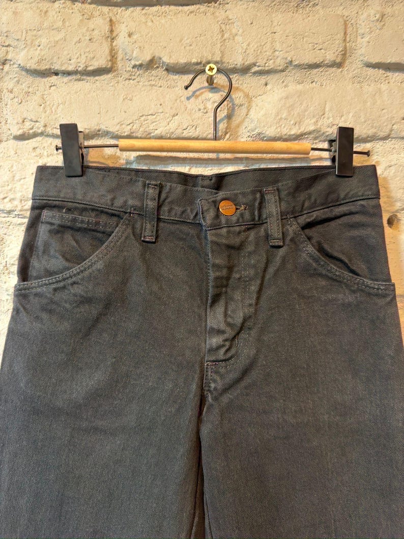 K&ouml;nnte beinhalten: Nahaufnahme einer anthrazitfarbenen Jeanshose, die an einem Holzb&uuml;gel h&auml;ngt. Die Hose hat einen Knopfverschluss und Vordertaschen. Der Hintergrund ist eine wei&szlig;e Backsteinmauer.