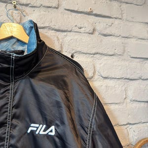 Op de afbeelding: Zwarte en lichtblauwe FILA-jas met witte stiksels en het FILA-logo op de borst. De jas hangt aan een houten hanger tegen een witte bakstenen muur.