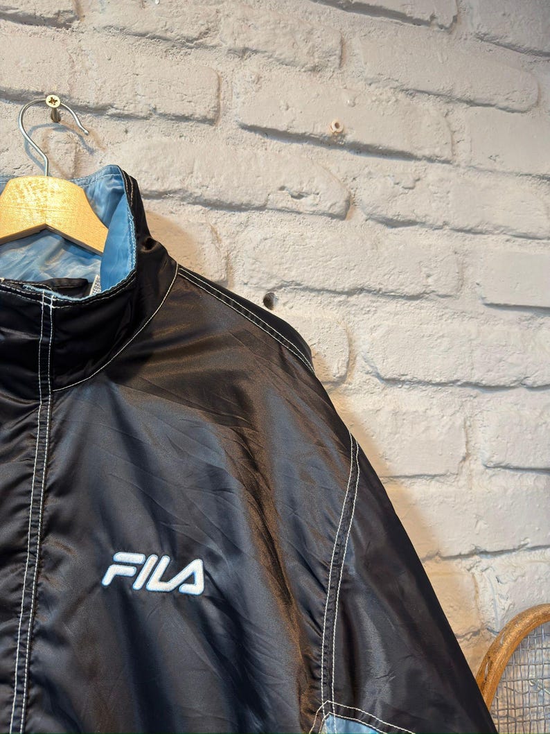 Op de afbeelding: Zwarte en lichtblauwe FILA-jas met witte stiksels en het FILA-logo op de borst. De jas hangt aan een houten hanger tegen een witte bakstenen muur.