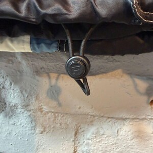 Op de afbeelding: Close-up van een zwarte jas met witte stiksels en een trekkoordsluiting. Het trekkoord heeft een zwarte plastic toggle met het merklogo. De jas heeft een blauw en wit accent.