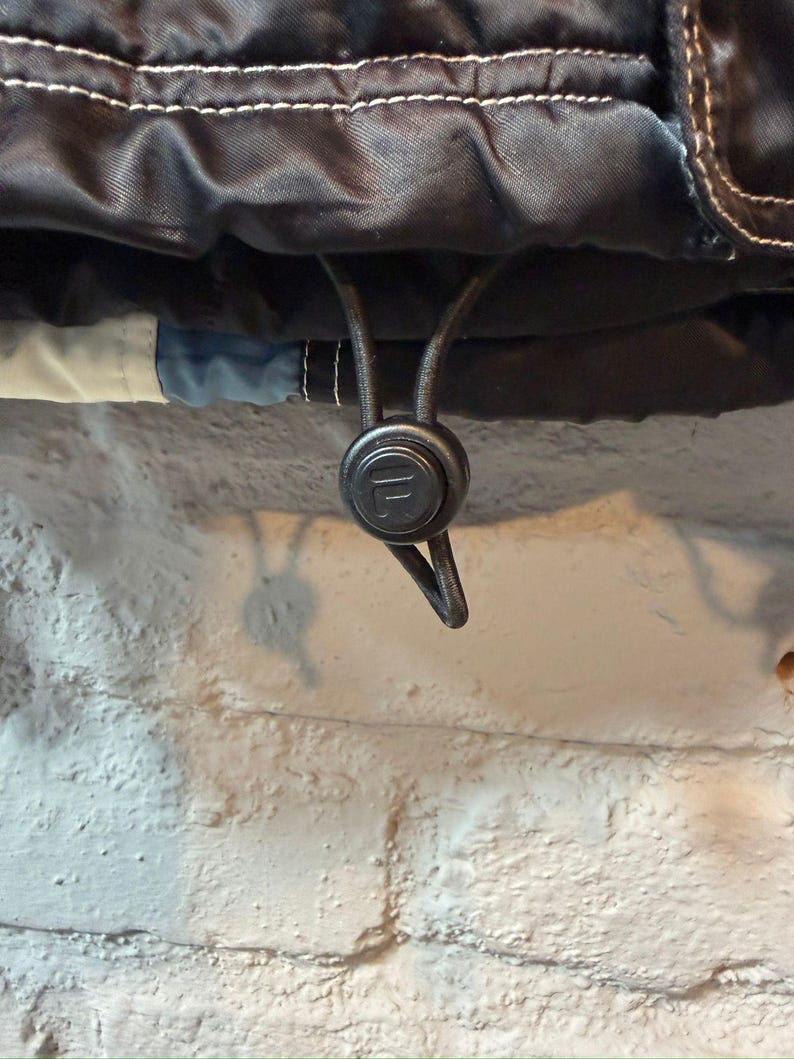 Op de afbeelding: Close-up van een zwarte jas met witte stiksels en een trekkoordsluiting. Het trekkoord heeft een zwarte plastic toggle met het merklogo. De jas heeft een blauw en wit accent.