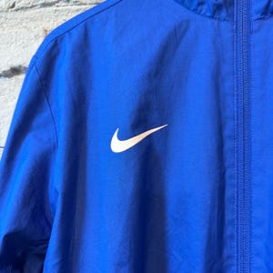 Puede incluir: Chaqueta deportiva azul real con el logotipo blanco de Nike en la parte delantera. La chaqueta tiene cremallera completa y capucha. El material parece ser una tela ligera y resistente al agua.