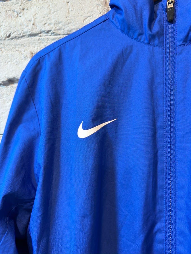 Puede incluir: Chaqueta deportiva azul real con el logotipo blanco de Nike en la parte delantera. La chaqueta tiene cremallera completa y capucha. El material parece ser una tela ligera y resistente al agua.