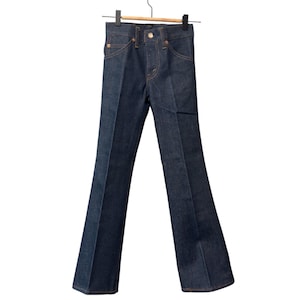 Può includere: Jeans in denim blu scuro con gamba a zampa, appesi a una gruccia di legno. I jeans hanno il classico stile a cinque tasche, una chiusura con bottone e cerniera e rivetti color rame. I jeans sono a vita media e sembrano mai indossati.