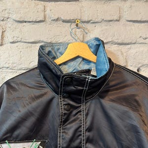 Op de afbeelding: Zwarte FILA-jas met een lichtblauwe kraagvoering en witte stiksels. De jas heeft een knoopsluiting en een patch met een berenafbeelding en de tekst "EHC Neuwied". De jas hangt aan een houten kleerhanger.