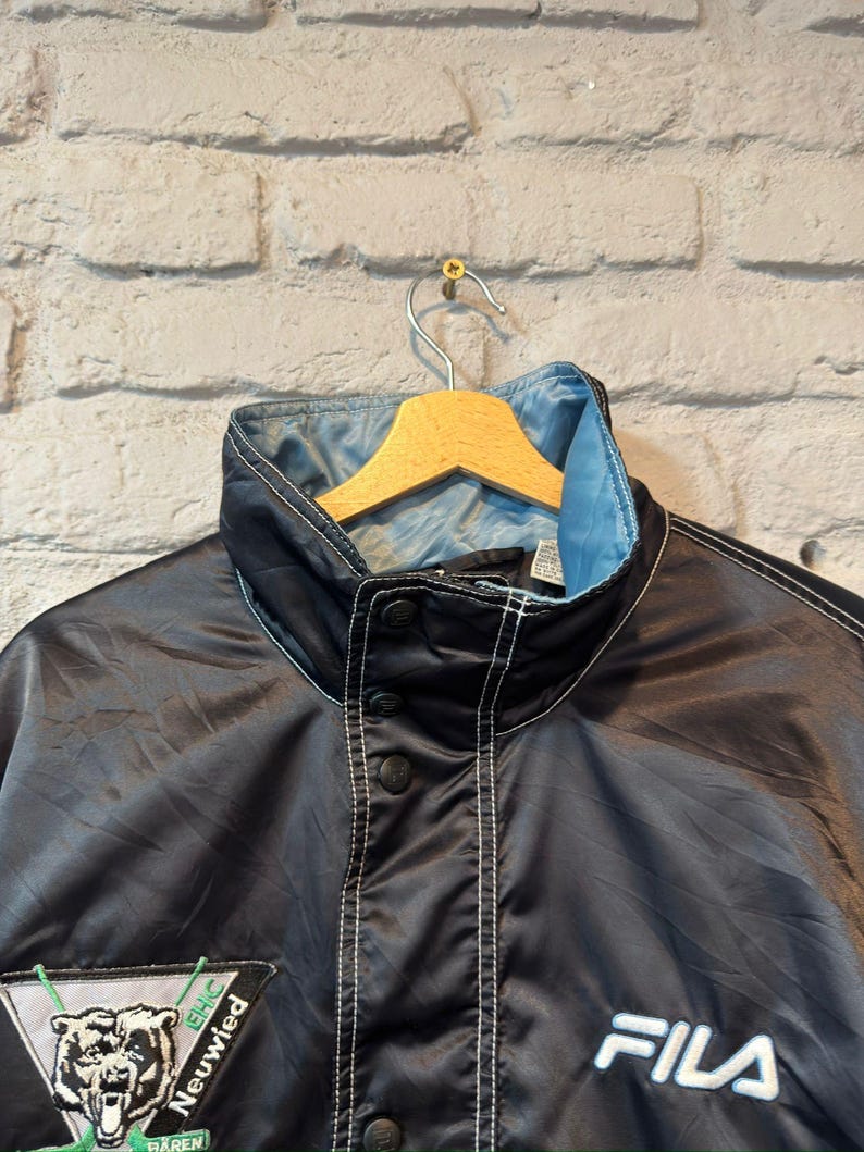 Op de afbeelding: Zwarte FILA-jas met een lichtblauwe kraagvoering en witte stiksels. De jas heeft een knoopsluiting en een patch met een berenafbeelding en de tekst "EHC Neuwied". De jas hangt aan een houten kleerhanger.