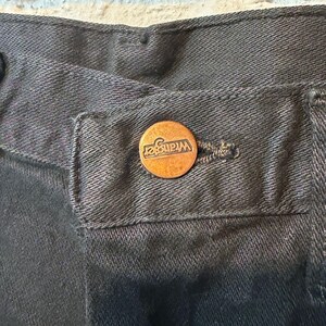 K&ouml;nnte beinhalten: Nahaufnahme einer schwarzen Jeans aus Denim mit einem kupferfarbenen Knopf. Der Knopf ist mit dem Markennamen "Wrangler" gepr&auml;gt. Die Jeans hat ein klassisches F&uuml;nf-Taschen-Design und einen Knopfverschluss.