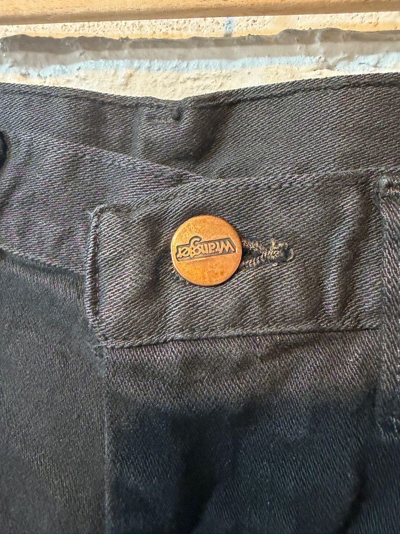K&ouml;nnte beinhalten: Nahaufnahme einer schwarzen Jeans aus Denim mit einem kupferfarbenen Knopf. Der Knopf ist mit dem Markennamen "Wrangler" gepr&auml;gt. Die Jeans hat ein klassisches F&uuml;nf-Taschen-Design und einen Knopfverschluss.