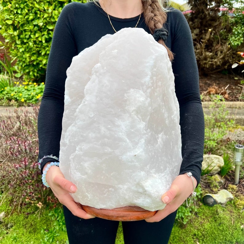 May include: A large, pale pink quartz crystal on a round, brown wooden base. The crystal has a rough, textured surface. The background features green foliage. The crystal is held by a person wearing a black long-sleeved shirt.