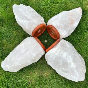May include: Four large, white Himalayan salt lamps with a natural, rough texture. Each lamp has a round, reddish-brown wooden base. The lamps are arranged in a flower-like pattern on green grass.