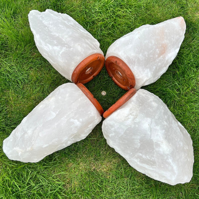May include: Four large, white Himalayan salt lamps with a natural, rough texture. Each lamp has a round, reddish-brown wooden base. The lamps are arranged in a flower-like pattern on green grass.