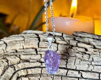 May include: A silver chain necklace with a raw, purple amethyst pendant. The necklace is displayed on a piece of weathered wood, with a lit candle in the background, creating a warm, natural aesthetic.