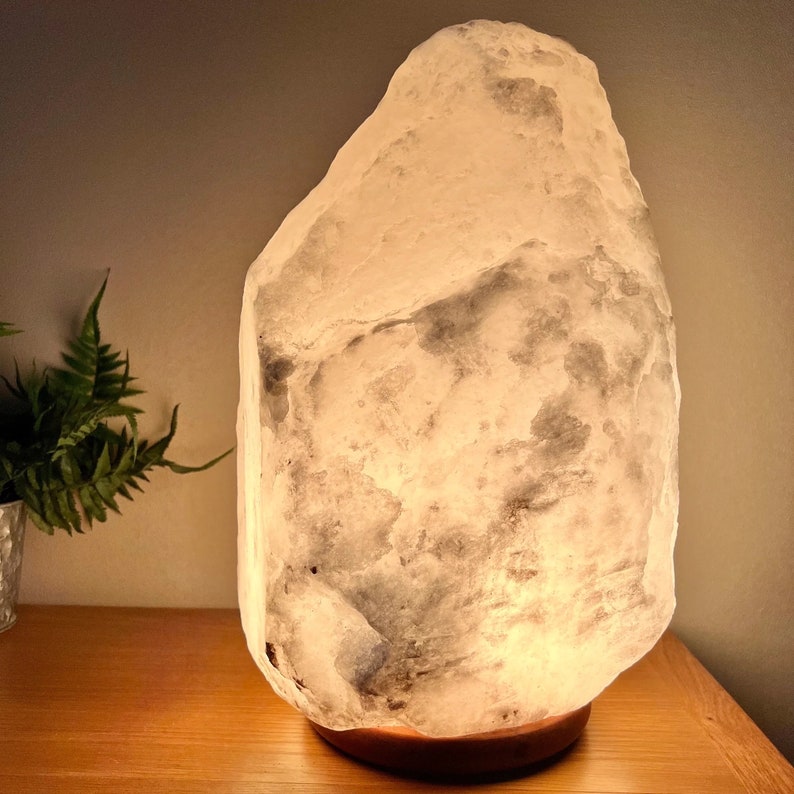 May include: A large, illuminated salt lamp with a rough, textured surface. The lamp emits a warm, soft glow, and sits on a wooden base. A small potted plant is visible to the left of the lamp.