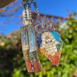 May include: Two crystal keychains with silver chains. One keychain features a clear crystal with embedded colourful stones and the other features a clear crystal with a silver Hamsa hand charm and embedded colourful stones.