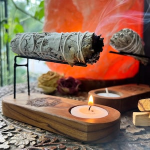 May include: A wooden stand with two candle holders and a metal holder for burning sage. The stand has a yin yang symbol carved into it. A sage stick is burning in the holder, with smoke rising from it.
