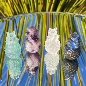 May include: Four carved gemstone pineapple figurines in shades of green, purple, clear, and dark gray. The pineapples are displayed on a reflective surface with a blurred background of green and yellow leaves. Each pineapple has a unique color and texture.