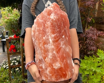 Könnte beinhalten: Eine große, orange-rosa Himalaya-Salzlampe mit einem Holzsockel. Die Lampe wird von einer Person gehalten.