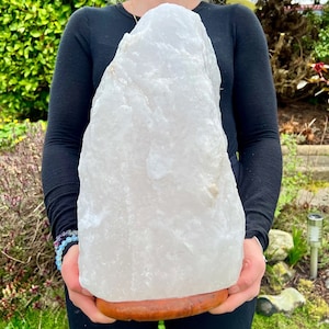 May include: A large, white crystal rock lamp on a wooden base. The crystal has a rough, natural texture and is held up by a brown, circular wooden base. The lamp is being held in front of a green background.