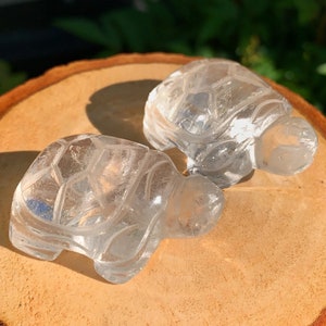May include: Two clear quartz crystal turtle figurines on a wooden surface. The turtles are carved with detailed shell patterns.