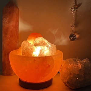 以下が含まれることがあります： 温かいオレンジ色の光を放つヒマラヤ岩塩ランプ。大きな岩塩の結晶で満たされたボウルが特徴です。ランプは点灯しており、周囲に柔らかな光を投げかけています。