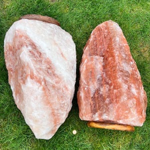 May include: Two large Himalayan salt rocks, one is a light pink color and the other is a darker orange color. Both rocks are sitting on a wooden base.