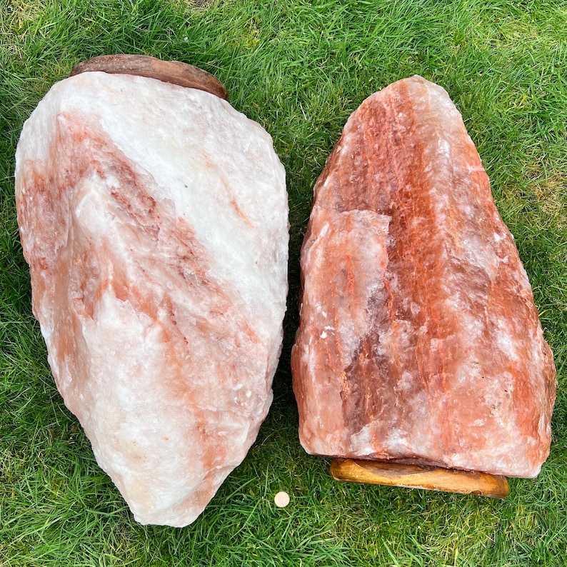 May include: Two large Himalayan salt rocks, one is a light pink color and the other is a darker orange color. Both rocks are sitting on a wooden base.