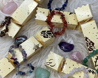 Può includere: Una collezione di braccialetti di pietre preziose colorate esposti su scatole di cracker di Natale dorate e bianche. I braccialetti sono realizzati con diverse pietre, tra cui ametista, corniola e quarzo rosa. I cracker sono decorati con stelle dorate e fiocchi di neve.