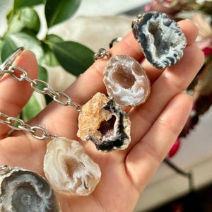 Könnte beinhalten: Eine Sammlung von Geoden-Anhängern an einer silbernen Kette. Die Geoden weisen verschiedene Farben auf, darunter Weiß, Braun und Grau, mit sichtbaren Kristallformationen. Die Anhänger werden in einer Hand gehalten.