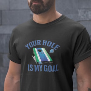 以下が含まれることがあります： 青い文字で「Your Hole Is My Goal」と書かれた、白と緑のコー​​ンホールボードのグラフィックが描かれた黒いTシャツです。