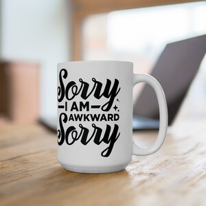 May include: White ceramic coffee mug with a black design that reads "Sorry - I am - Awkward Sorry".