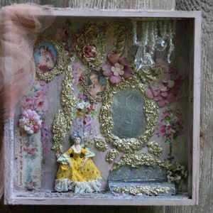 Può includere: Una scatola a ombre di ispirazione vintage con una bambola di porcellana vestita con un abito giallo e pizzo. La scatola è decorata con fiori, dettagli dorati e uno specchio piccolo. Il testo "Forever wait of dawn will all"