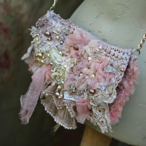 May include: A delicate, crocheted handbag with a chain strap. The bag is adorned with layers of lace, ruffles, and pearls in shades of pink and white.