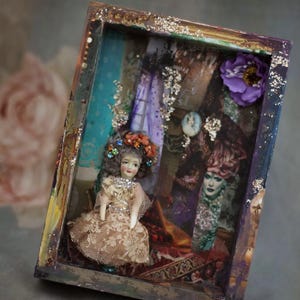 May include: A vintage doll wearing a lace dress and a flower crown sits in a shadow box with a colorful background. The box is decorated with glitter and other embellishments.
