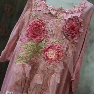 May include: A light pink tunic top with long sleeves, adorned with fabric flowers, lace, and bead embellishments. The neckline is trimmed with lace and pearls. The floral details are in shades of pink and peach, with green leaf accents.