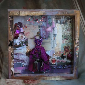 May include: A framed shadow box art piece with a purple dress and a variety of textures and colors. The dress is adorned with purple beads and has a black bow. The background is a collage of vintage papers and fabrics.