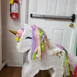 May include: A white unicorn pinata with a gold horn, pink and yellow flowers, and a rainbow tail. The pinata is standing on a wooden floor.