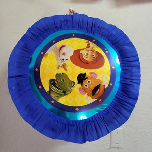 May include: A blue and yellow round pinata with a blue ruffled border. The pinata features a cartoon image of characters from the Toy Story movie franchise. The characters include Woody, Mr. Potato Head, Rex the dinosaur, and a unicorn.