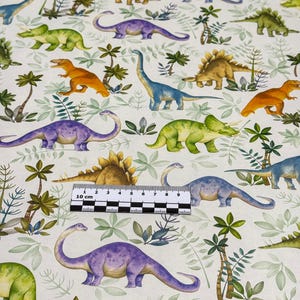 Pode incluir: Um tecido branco com um padrão repetido de dinossauros coloridos e palmeiras verdes. O tecido é medido com uma régua em centímetros.