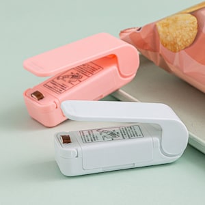Könnte beinhalten: Zwei handgehaltene Heißsiegler, einer rosa und einer weiß, zum Verschließen von Plastiktüten. Die Siegelgeräte haben einen Griff, der sich zum Aktivieren des Heizelements herunterklappen lässt.