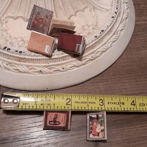 May include: Miniature fairy tale books, including "Snow White" and "Alice in Wonderland," are displayed. The books have aged, vintage covers with detailed illustrations and text. A measuring tape shows the books are approximately 1 inch in length.