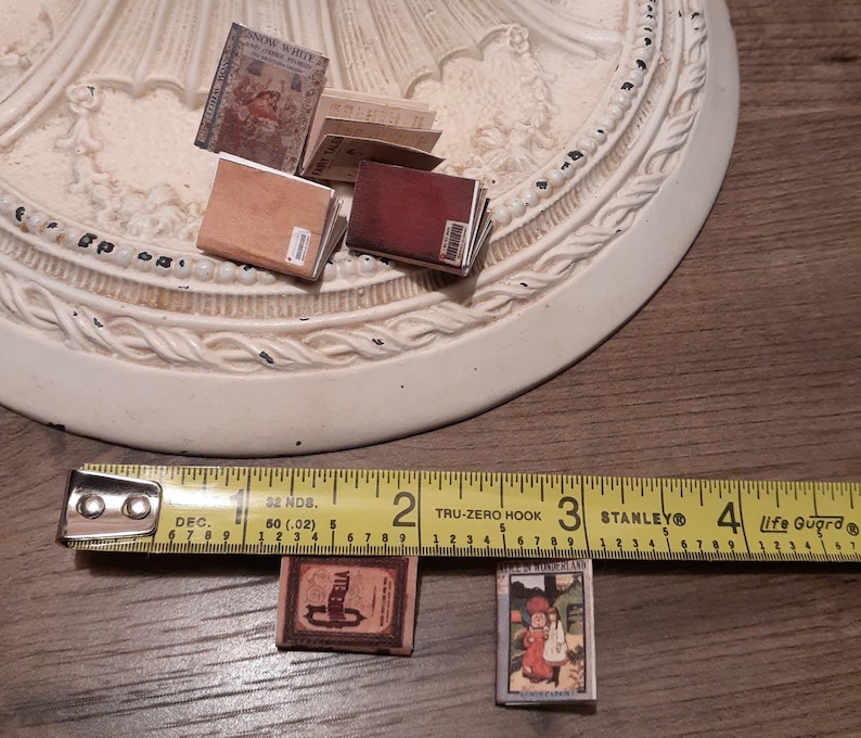 May include: Miniature fairy tale books, including "Snow White" and "Alice in Wonderland," are displayed. The books have aged, vintage covers with detailed illustrations and text. A measuring tape shows the books are approximately 1 inch in length.