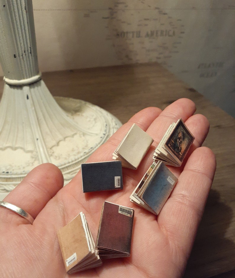 May include: A collection of miniature books in various colors, including tan, navy, and burgundy, are held in a hand. The books have white pages and some feature small illustrations. Each book has a small white label.