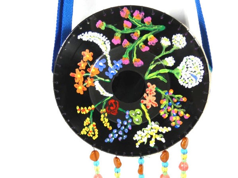 May include: A black vinyl record painted with a colorful floral design. The record is hanging from a blue ribbon and has colorful beads hanging from the bottom.
